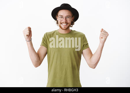 Yes we did it. Portrait of emotional happy and delighted handsome young hipster bearded male in glasses and hat smiling broadly as shaking clenched Stock Photo