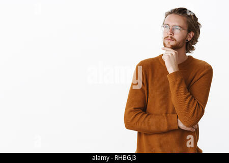 Waist-up shot of romantic and thoughtful male writer in glasses and earrings touching beard squinting at looking focused at upper left corner Stock Photo