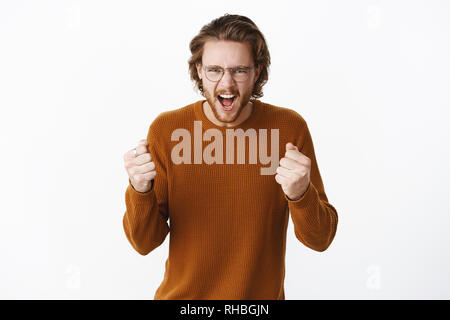 Studio shot of expressive good-looking european bearded guy in sweater and glasses yelling with fure and rage clenching raised fists as cheering and Stock Photo