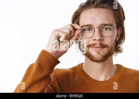 Unaltered shot of handsome friendly-looking smart fair-haired guy in glasses and sweater touching rim of eyewear and smiling at camera, looking with Stock Photo