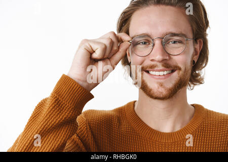 Close-up shot of satisfied accomplished and smart bearded ginger guy in sweater touching glasses and smiling broadly with friendly grin, standing Stock Photo