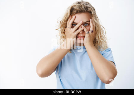 Girl peeking as not liking surprises. Cute and young attractive blonde with short haircut in blue t-shirt holding hands on face, cover sight and peek Stock Photo