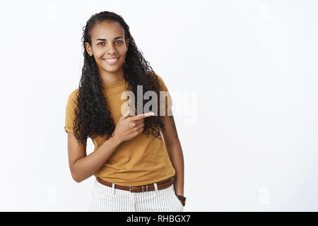 Studio shot of shy and cute pretty young african american woman with long curly hair bending as pointing right with index finger and smiling, holding Stock Photo