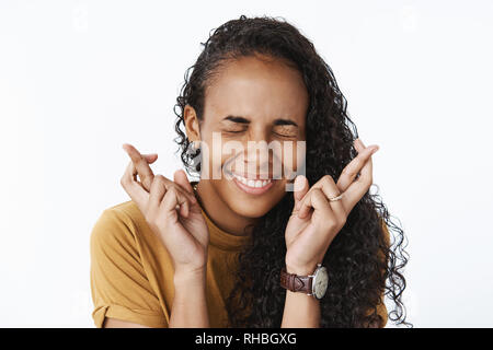 Close-up shot of happy excited and optimistic young african-american woman making wish with smile and closed eyes crossing fingers for good luck as Stock Photo
