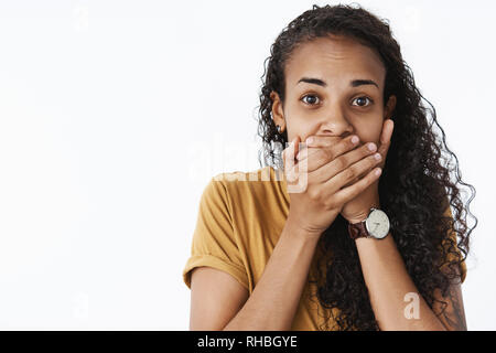 Close-up shot of shocked and speechless young impressed dark-skinned girl with curly hair closing mouth with palms as being astonished and sweep from Stock Photo