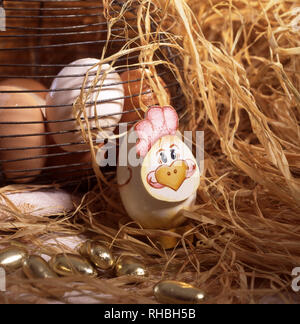Boiled egg with painted face for Easter set amongst straw. Basket of fresh eggs in the background. Stock Photo