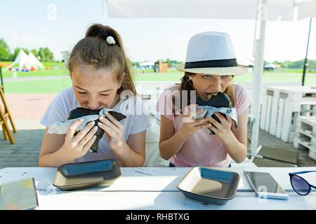 Teens girls with an appetite eats black fast food burger. Summer street cafe, recreation area, city park background Stock Photo