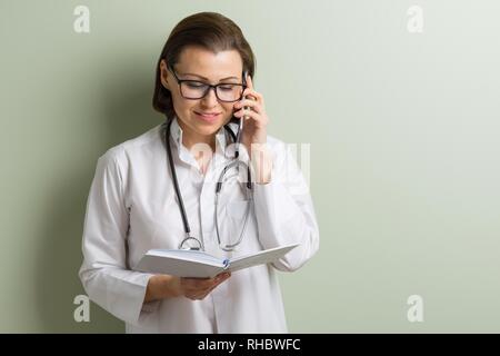 Medical doctor woman with stethoscope looks reading his work notebook talking mobile phone. Background green wall in the clinic, copy space. Stock Photo