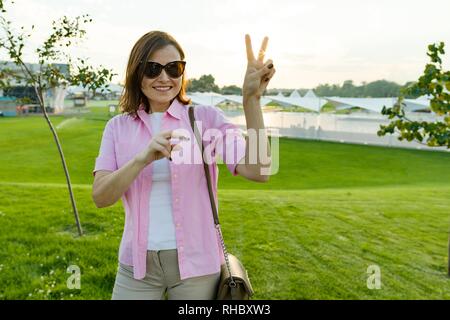 Middle-aged woman showing sign of victory, background horizon with sunset, green lawn Stock Photo