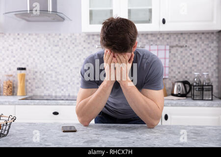 Frustrated Man Covering His Face With Two Hand Sitting In Kitchen Stock Photo
