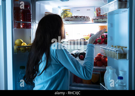 Rear View Of A Young Woman Standing In Front Of An Open Refrigerator Shrugging Stock Photo
