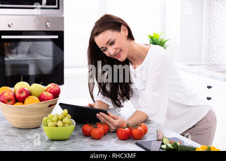 Happy Young Woman Using Digital Tablet Near Fresh Vegetables On Kitchen Counter Stock Photo