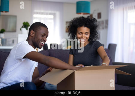 Young black couple open cardboard box unpacking sitting on couch Stock Photo