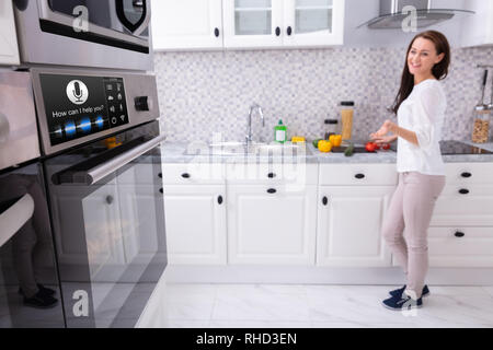 Happy Young Woman Looking At Oven With Voice Recognition Function In Kitchen Stock Photo