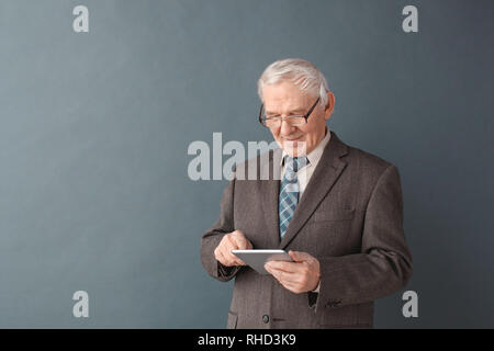 Senior male teacher wearing eyeglasses studio standing isolated on gray wall using digital tablet browsing internet concentrated Stock Photo