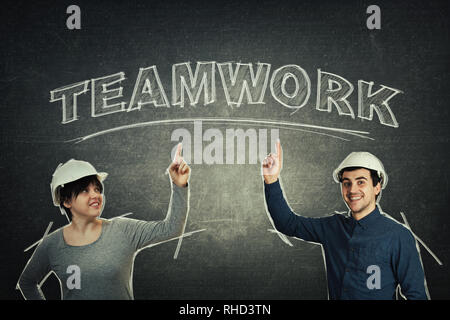 Positive thinking engineers wearing protective helmet index finger pointed showing chalk written teamwork text on blackboard. Sharing thoughts togethe Stock Photo