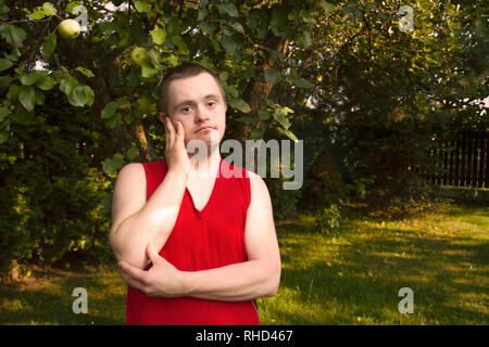 Portrait of young man with Down Syndrome standing in the garden under apple tree. Adult person with in red blouse in summer, green loan. Stock Photo