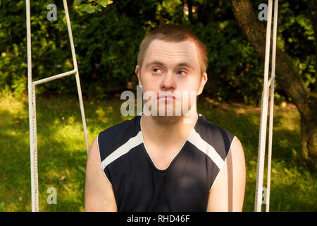 Portrait of young person with Down Syndrome sitting on a swing in the garden, backyard during summer day. Young handsome looks up looks up. Face close Stock Photo