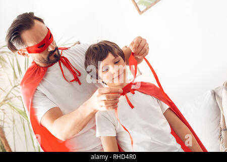 Father and little son wearing superheroe costumes together at home standing man healping boy to wear mask carefully Stock Photo