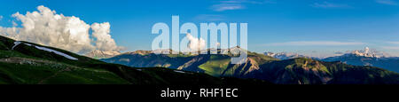 Wide mountain panorama.Green hills with snow on peaks in evening sunlight. Dark shadows. Blue sky with some clouds. Summer in Georgia. Stock Photo