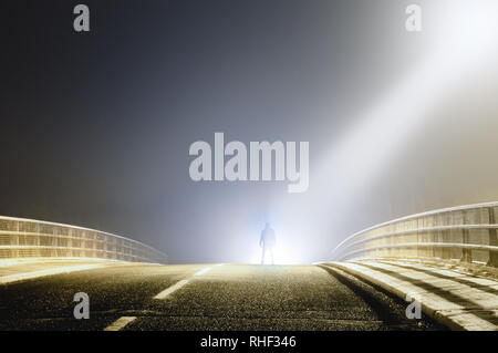 A sinister hooded figure silhoutted by a light beam from a UFO on a spooky misty road at night. Stock Photo