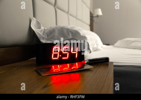 Rectangular alarm clock on bedside table with big red numbers, big kitchen knife in hotel room. Concept sleep disturbance, oversleep work, night shift Stock Photo