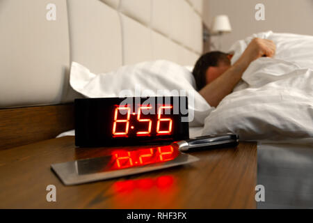 Alarm clock on the bedside table with red numbers, kitchen knife, sleeping man in bed in room. Concept chef sleeping after hard day at restaurant, sle Stock Photo