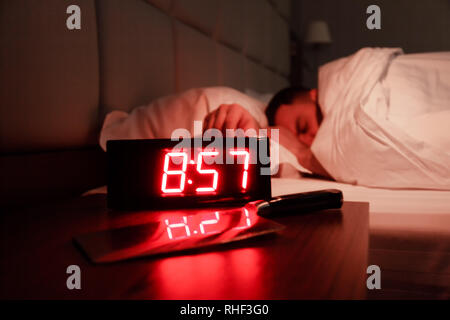 Alarm clock on the bedside table with red numbers, kitchen knife, sleeping man in bed in dark room. Concept chef sleeping after hard day at restaurant Stock Photo