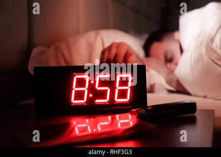 Alarm clock on the bedside table with red numbers, kitchen knife, sleeping man in bed in dark room. Concept chef sleeping after hard day at restaurant Stock Photo