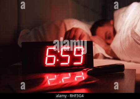 Alarm clock on the bedside table with red numbers, kitchen knife, sleeping man in bed in dark room. Concept chef sleeping after hard day at restaurant Stock Photo