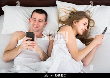 Young couple reading with smartphones turning their back away in bed at home Stock Photo