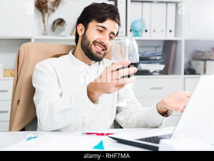 Cheerful brunette man holding glass of red wine and having video chat at office Stock Photo