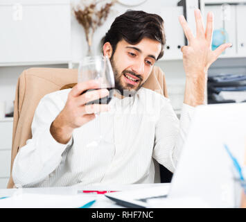 Cheerful brunette man holding glass of red wine and having video chat at office Stock Photo