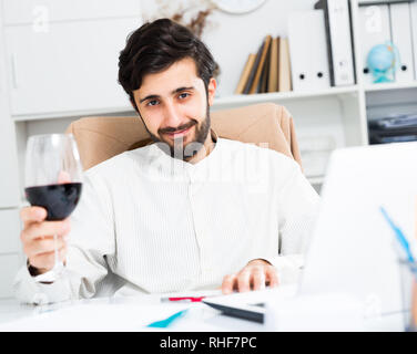 Handsome young office worker drinking red wine at office desk Stock Photo