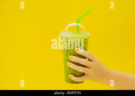 Green tea latte with ice in plastic cup and straw in female hand with yellow manicure on yellow background. Homemade Iced Matcha Latte Tea with Milk Stock Photo