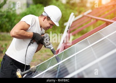 Profile view of technician connecting blue shiny solar photo voltaic panel to metal platform using electrical screwdriver on warm summer day. Stand-alone solar panel system installation concept. Stock Photo
