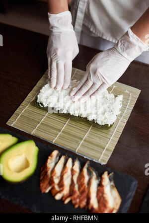 Close-up view of process of preparing rolling sushi. Nori and white rice on bamboo mat. Chef's hands touch rice. Chef starts cooking sushi. Eel and avocado Stock Photo