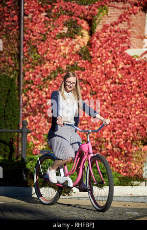 Young happy long-haired smiling blond woman in casual clothing and glasses cycling pink lady bicycle on bright warm sunny day on background of brick wall overgrown with beautiful red ivy leaves. Stock Photo