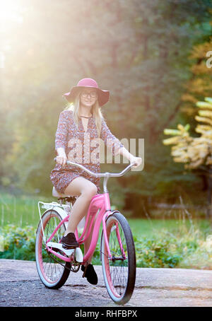Slim happy smiling blond fashionable attractive woman in glasses, short dress and red hat riding lady bicycle along paved park alley on beautiful green and golden trees lit by bright sun background. Stock Photo