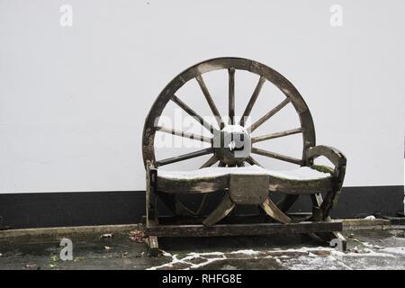 A natural wooden seat or bench for the garden, made from a wagon or mill wheel. It is covered lightly in snow and against a white wall ensuring the pa Stock Photo