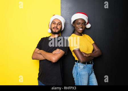 Young happy mixed race couple, indian man and african woman in santa hats standing on yellow black background Stock Photo