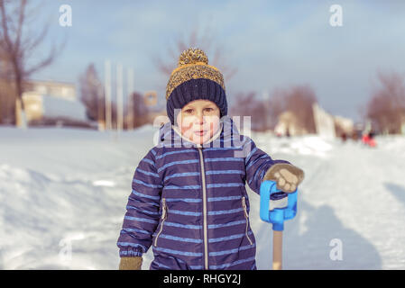 A little boy in the winter in city, in a blue jumpsuit and a warm yellow hat. In the hands of holding a spatula. Surprised resting in nature outside