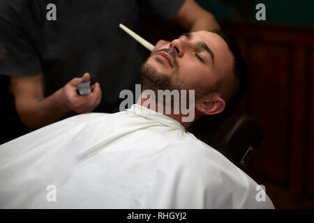 Close-up side view portrait of handsome young bearded Caucasian man getting beard grooming in modern barbershop Stock Photo