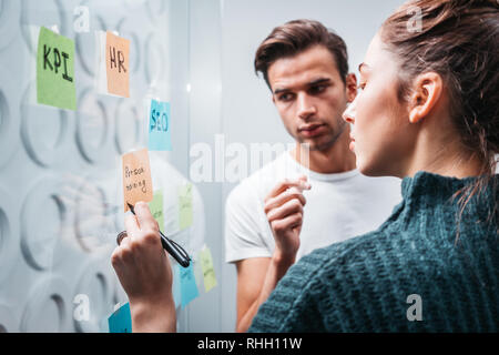 Team of project managers having meeting at modern office. Business colleagues discussing new ideas and using sticky glass note wall Stock Photo