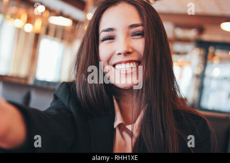 Beautiful charming brunette smiling asian girl taking selfie on frontal camera at cafe Stock Photo