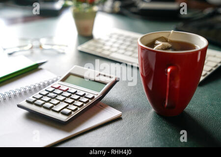 Work place with view of keyboard, notebook, old glasses , a red mug with tea on desk and main subject the calculator Stock Photo