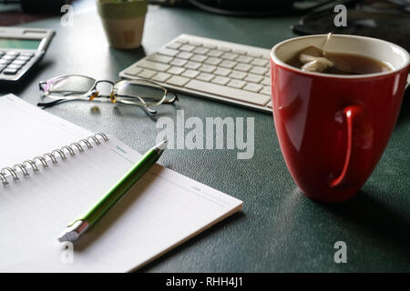Work place with view of keyboard, notebook, pen, old glasses and a red mug with tea on desk Stock Photo