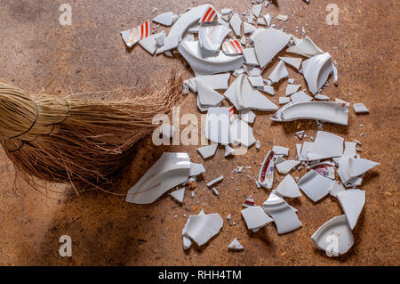 Straw broom sweeping pieces of shattered dishes. Splinters and fragments of the broken white ware on the floor. Metaphor of a family quarrel. Stock Photo