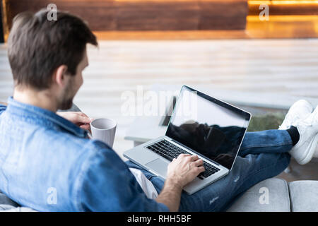 Young attractive smiling guy is browsing at his laptop, sitting at home on the gray sofa at home, wearing casual outfit. Stock Photo