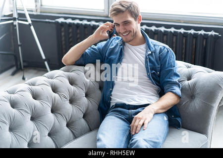 Young happy man sitting relaxed on sofa and talking on the phone. Stock Photo